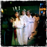 Elementary school girls dressed in white and the leader with candles for a crown walk towards the stage in a Santa Lucia performance.